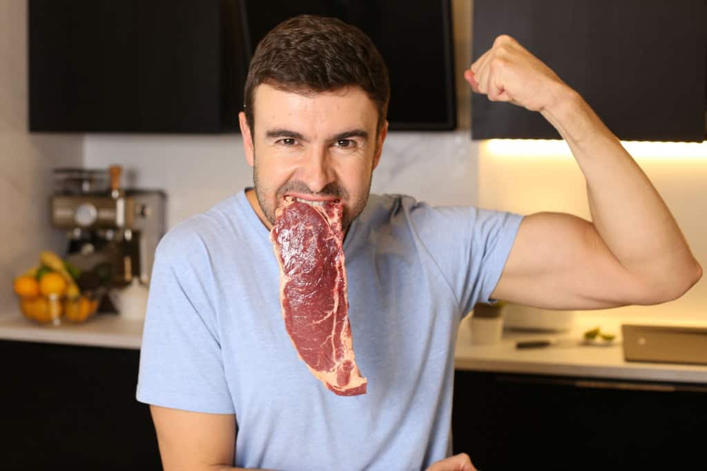 Homem com um bife cru na boca enquanto mostra os músculos do braço