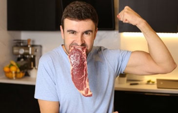 Homem com um bife cru na boca enquanto mostra os músculos do braço