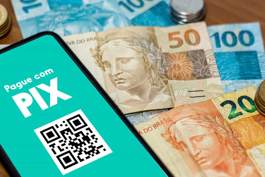 Smartphone com os dizeres "Pague com PIX" e um QR Code em cima de várias notas de real