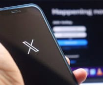 Ao fundo, desfocado, página de login do X; à frente, logo do X em um smartphone