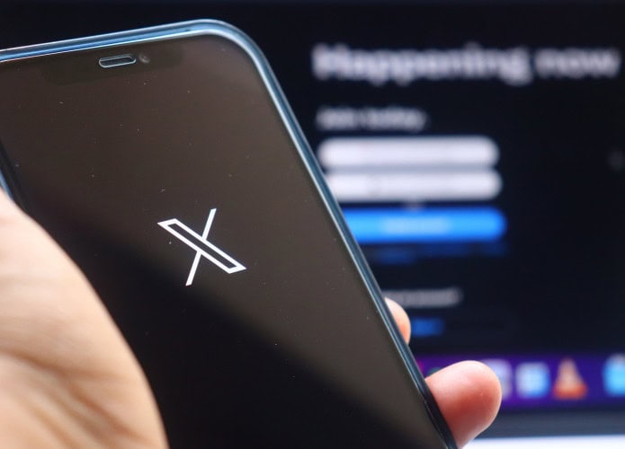 Ao fundo, desfocado, página de login do X; à frente, logo do X em um smartphone