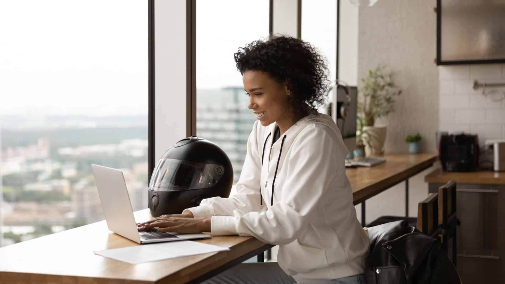 Empresária afro-americana sorrindo, usando laptop, trabalhando em projeto online em casa, sentada à mesa de madeira com capacete de motocicleta, jovem conversando ou comprando online, navegando em aplicativos