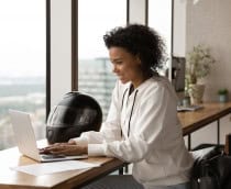 Empresária afro-americana sorrindo, usando laptop, trabalhando em projeto online em casa, sentada à mesa de madeira com capacete de motocicleta, jovem conversando ou comprando online, navegando em aplicativos