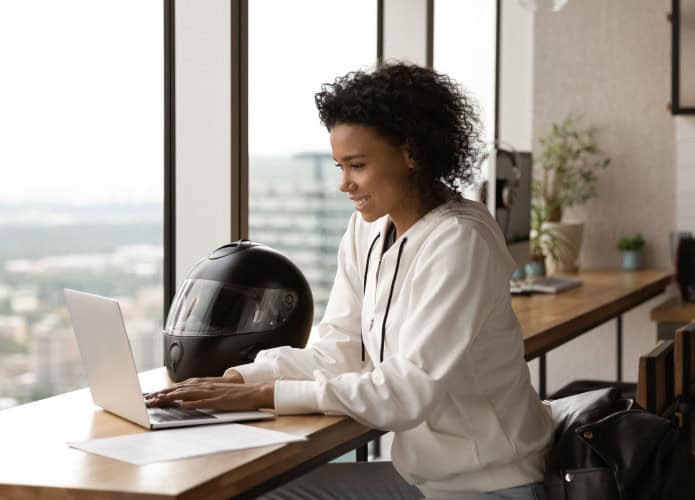 Empresária afro-americana sorrindo, usando laptop, trabalhando em projeto online em casa, sentada à mesa de madeira com capacete de motocicleta, jovem conversando ou comprando online, navegando em aplicativos