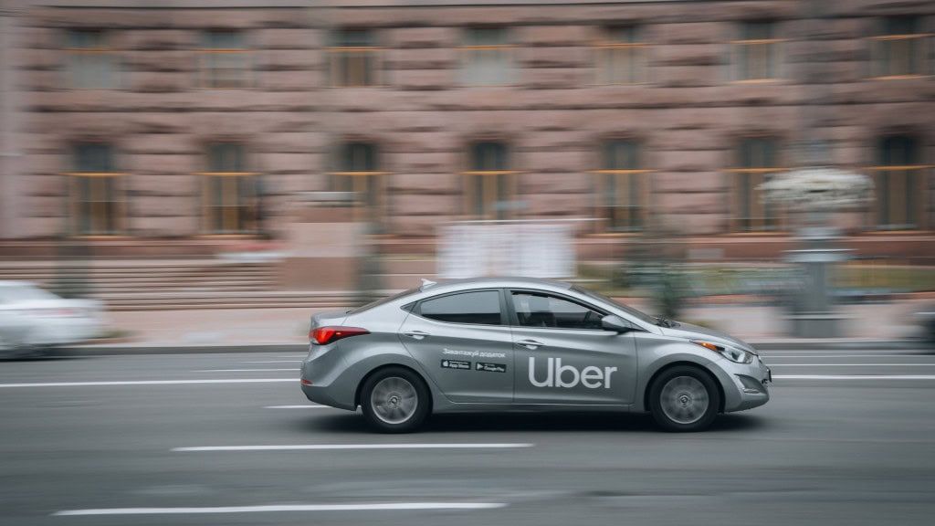 Carro cinza com logo da Uber em destaque cruza uma rua urbana, em movimento rápido diante de prédios desfocados ao fundo.