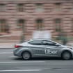 Carro cinza com logo da Uber em destaque cruza uma rua urbana, em movimento rápido diante de prédios desfocados ao fundo.