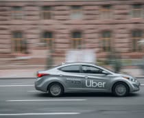 Carro cinza com logo da Uber em destaque cruza uma rua urbana, em movimento rápido diante de prédios desfocados ao fundo.