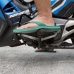 A imagem mostra o pé de uma pessoa usando chinelo verde sobre os pedais de uma motocicleta azul. O condutor parece prestes a trocar de marcha, com a moto estacionada sobre piso de concreto.