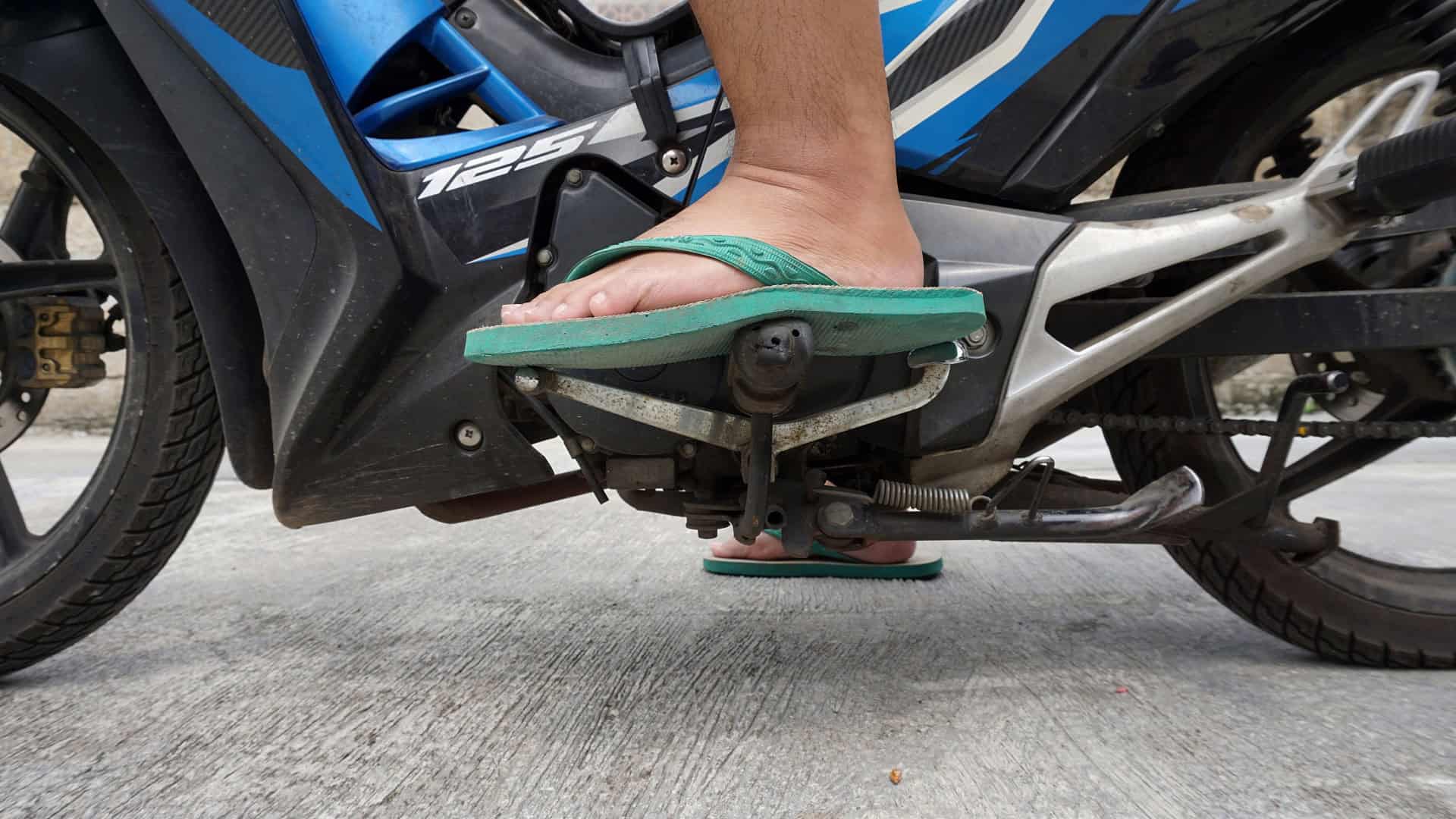 A imagem mostra o pé de uma pessoa usando chinelo verde sobre os pedais de uma motocicleta azul. O condutor parece prestes a trocar de marcha, com a moto estacionada sobre piso de concreto.