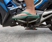 A imagem mostra o pé de uma pessoa usando chinelo verde sobre os pedais de uma motocicleta azul. O condutor parece prestes a trocar de marcha, com a moto estacionada sobre piso de concreto.