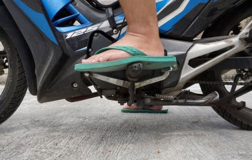 A imagem mostra o pé de uma pessoa usando chinelo verde sobre os pedais de uma motocicleta azul. O condutor parece prestes a trocar de marcha, com a moto estacionada sobre piso de concreto.
