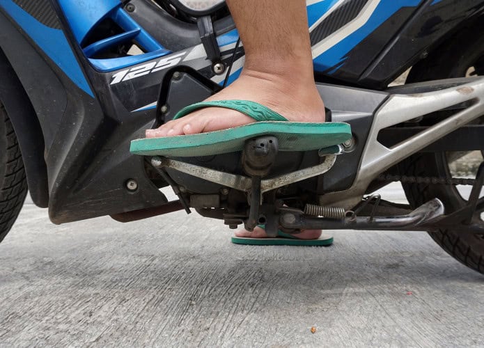 A imagem mostra o pé de uma pessoa usando chinelo verde sobre os pedais de uma motocicleta azul. O condutor parece prestes a trocar de marcha, com a moto estacionada sobre piso de concreto.