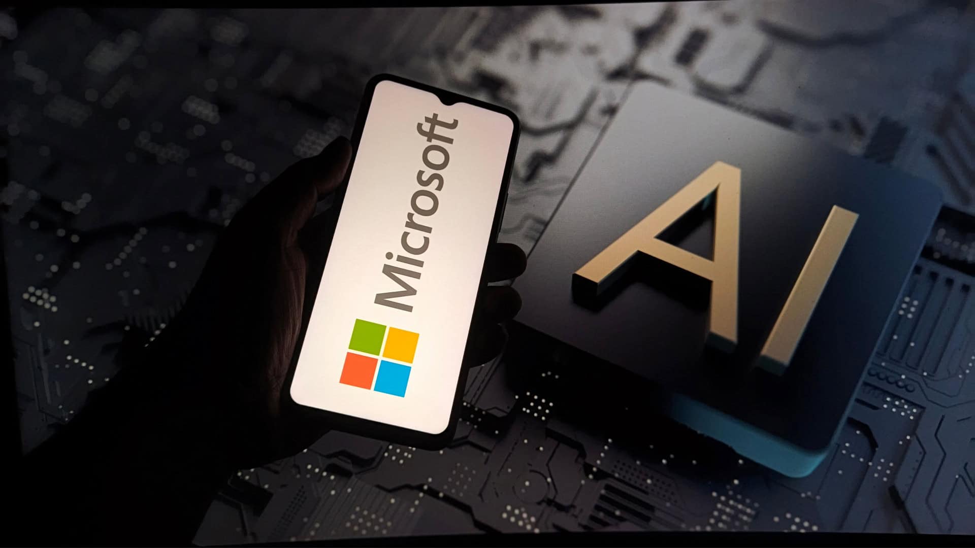 Pessoa segurando celular com logotipo da Microsoft na tela na frente de outra tela, esta grande, exibindo ilustração de chip de inteligência artificial (IA)