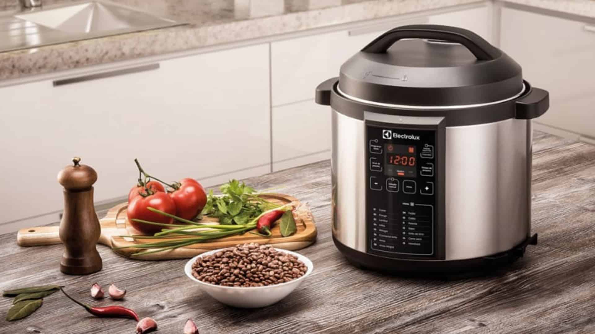 Panela de pressão elétrica em cima da mesa, ao lado de uma tábua de legumes e um moedor de sal