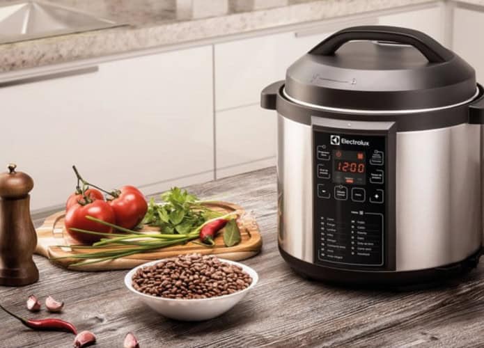 Panela de pressão elétrica em cima da mesa, ao lado de uma tábua de legumes e um moedor de sal