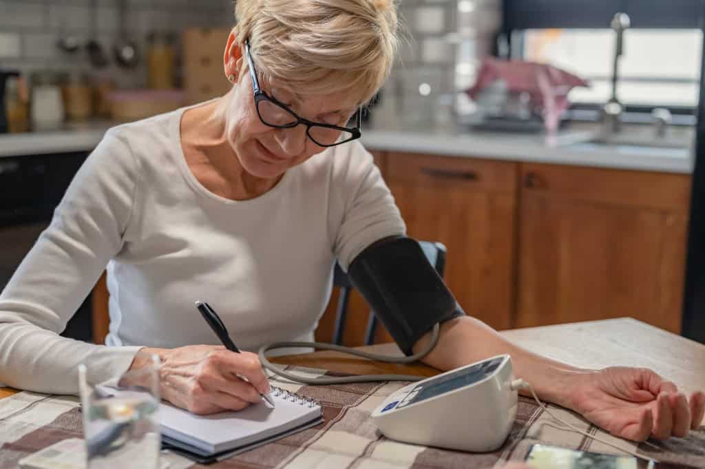 Uma senhora idosa mede sua pressão em aparelho digital em casa e anota resultados.