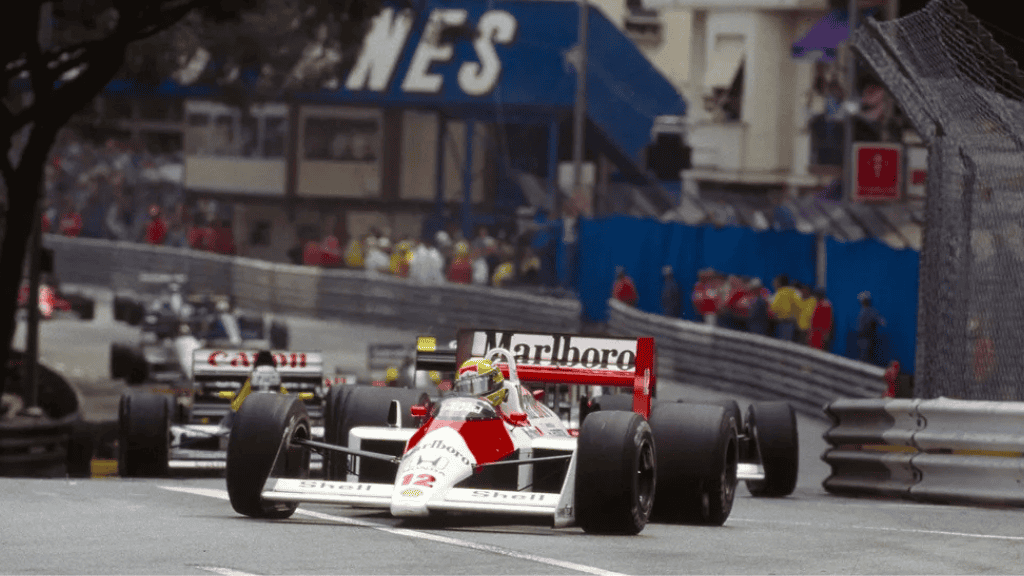Carro de Fórmula 1 da McLaren, pilotado por Ayrton Senna, lidera corrida nas ruas estreitas do GP de Mônaco, seguido por outros carros.