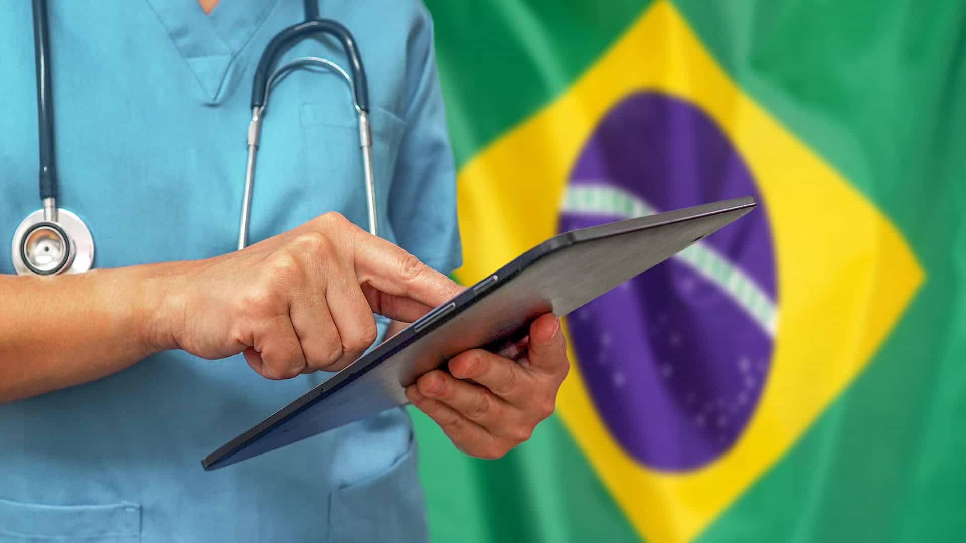 imagem mostra um médico mexendo em um tablet em frente a uma bandeira do brasil