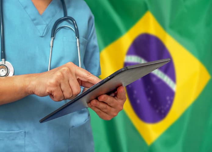 imagem mostra um médico mexendo em um tablet em frente a uma bandeira do brasil