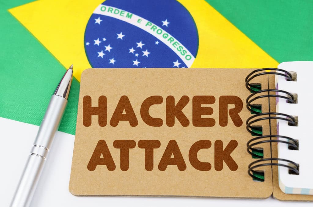 Ao fundo, uma bandeira do Brasil em cima de uma mesa branca; em cima dela, uma caneta e um bloco de notas; No bloco de notas, lê-se "hacker attack"