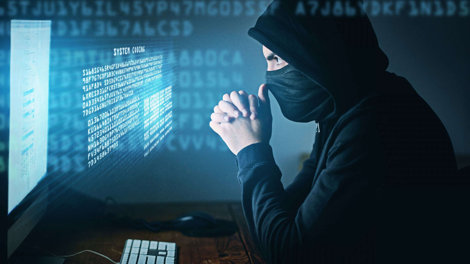 Pessoa de capuz e máscara com as mãos na região do nariz olhando para uma tela de computador