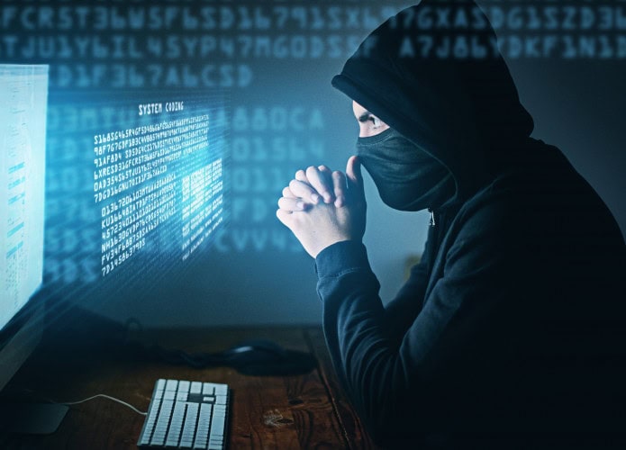 Pessoa de capuz e máscara com as mãos na região do nariz olhando para uma tela de computador