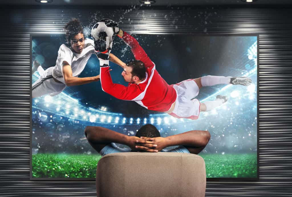 Imagem mostra telespectador sentado em poltrona e assistindo a imagem dinâmica dos jogadores em movimento e a clareza da cena de futebo