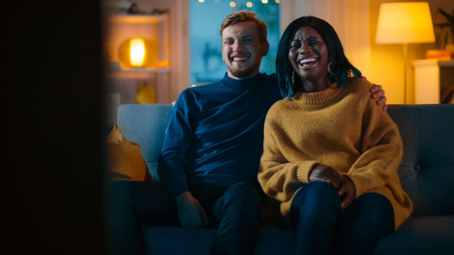 Casal jovem e diverso assistindo comédia na TV, rindo e aproveitando o momento no sofá / Crédito: Gorodenkoff – Shutterstock (divulgação)