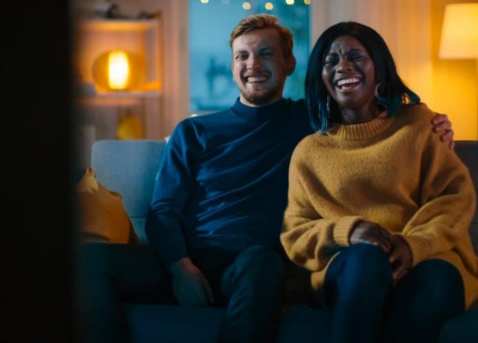 Casal jovem e diverso assistindo comédia na TV, rindo e aproveitando o momento no sofá / Crédito: Gorodenkoff – Shutterstock (divulgação)