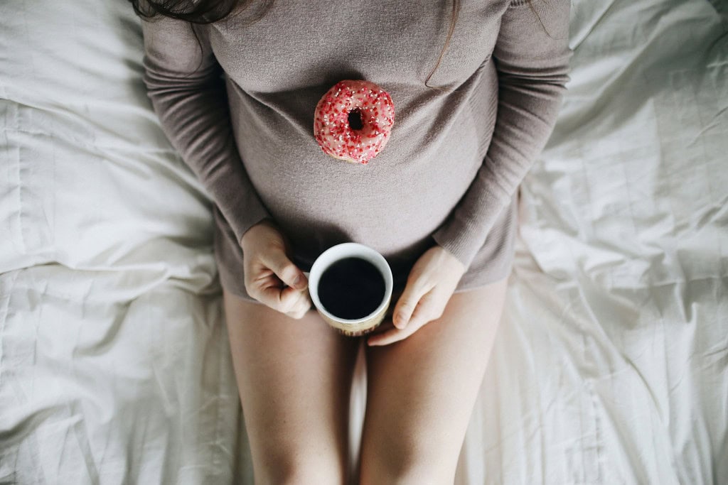 Imagem mostra corpo de grávida deitada na cama com um donut em cima da barriga e segurando uma xícara de café