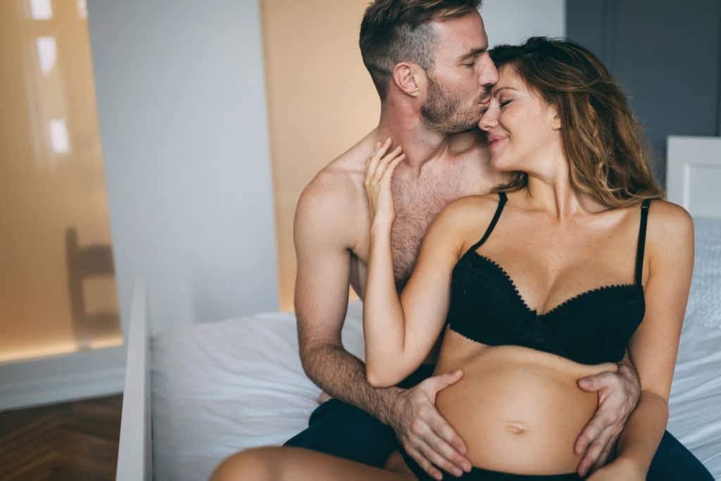 Imagem mostra momento de intimidade entre casal, com homem beijando a testa da mulher grávida e com as mãos na barriga dela