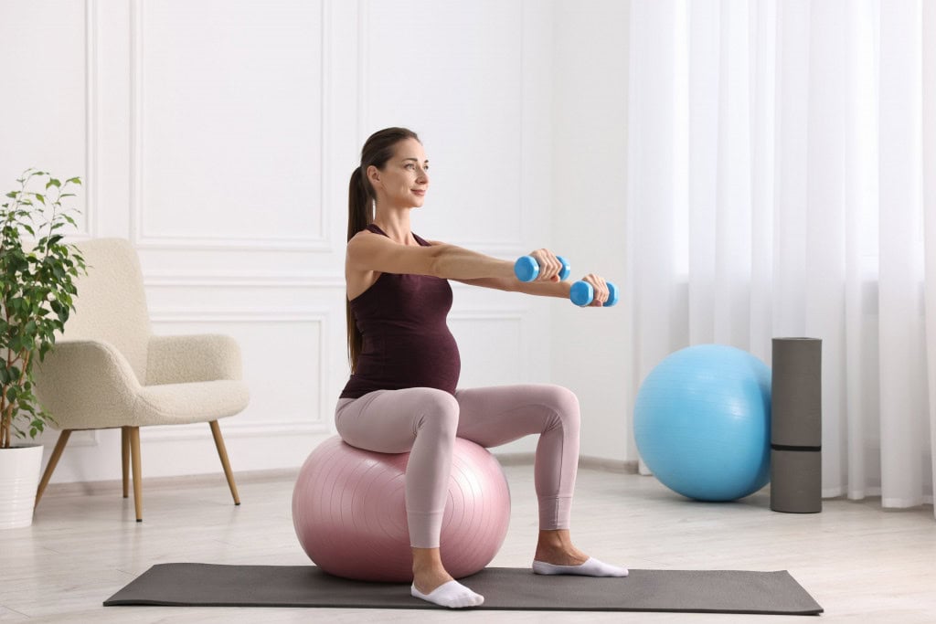 Grávida praticando atividade física, sentada em bola de pilates e com halteres nas mãos