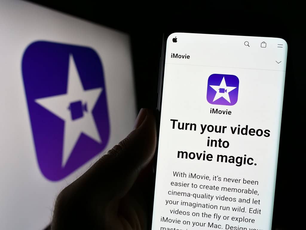 Imagem mostra pessoa segurando um celular com o site do software de edição de vídeo iMovie (Apple Inc.) na tela, em frente ao logotipo