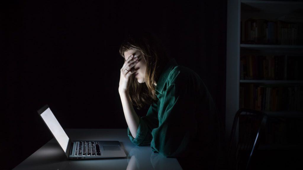 Mulher jovem cansada e exausta esconde os olhos com a mão enquanto usa o laptop à noite.