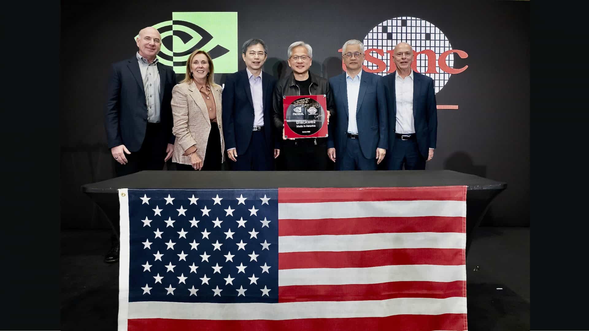 Jensen Huang e outros representantes de Nvidia e TSMC atrás de uma mesa com a bandeira dos EUA; atrás deles, um cartaz com os logos das duas empresas