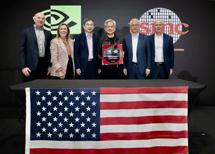 Jensen Huang e outros representantes de Nvidia e TSMC atrás de uma mesa com a bandeira dos EUA; atrás deles, um cartaz com os logos das duas empresas