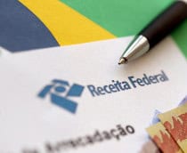 Ao fundo, bandeira do Brasil; em cima, um papel com o logo da receita federal, uma caneta e algumas notas de real