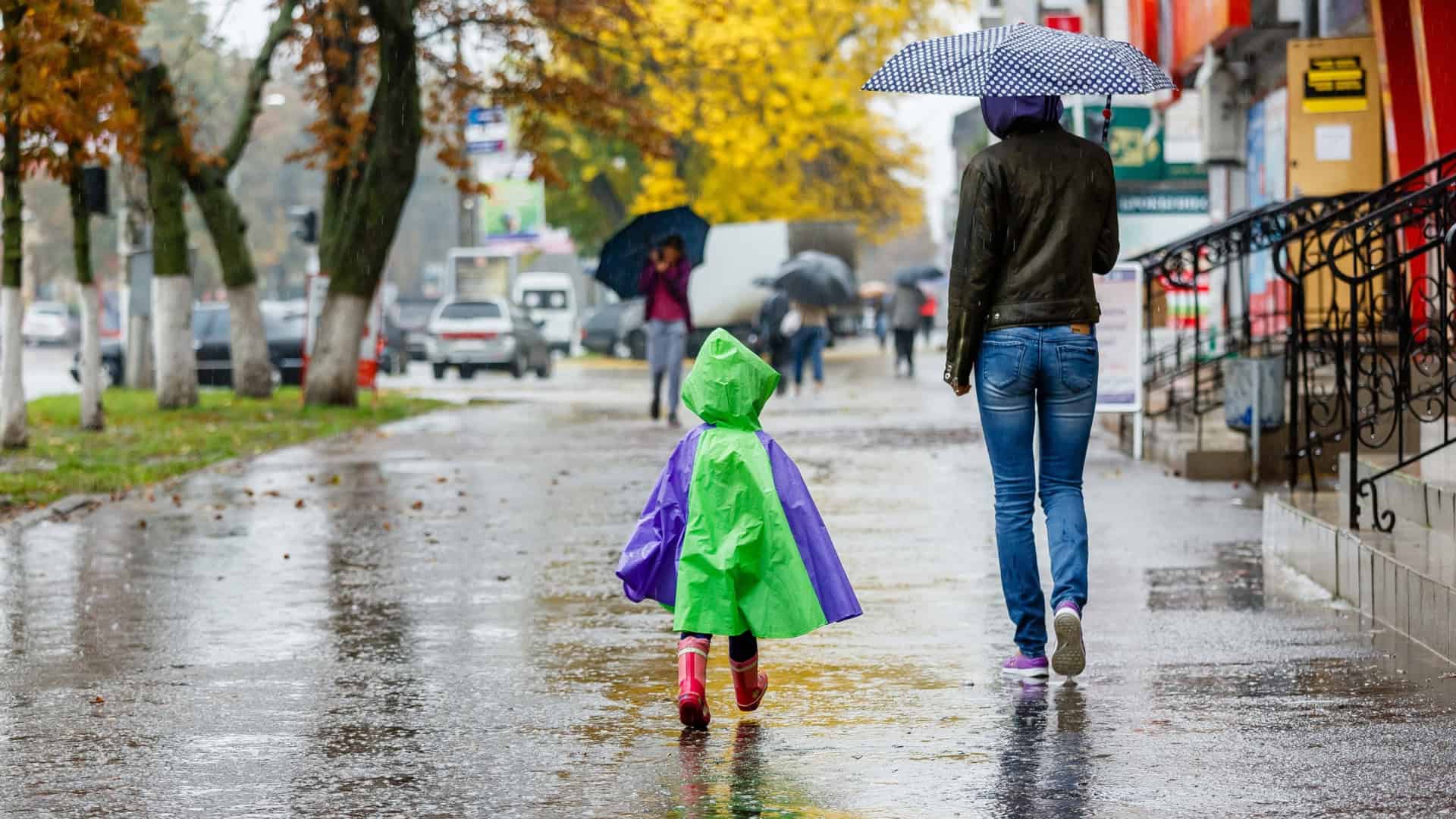 Mulher e criança de costas andando na chuva; a mulher está segurando um guarda-chuva