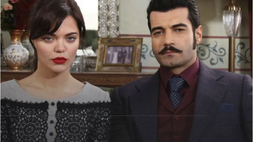 Duas pessoas estão sentadas lado a lado em um ambiente elegante e antigo; uma mulher com batom vermelho e blusa clara rendada, e um homem de terno escuro com camisa vinho. Atrás deles há flores, espelho e uma foto emoldurada, sugerindo um cenário clássico.