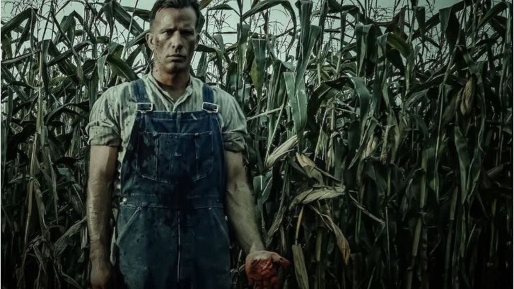 Homem com macacão e camisa, saindo do milharal com as mãos cheias de sangue.