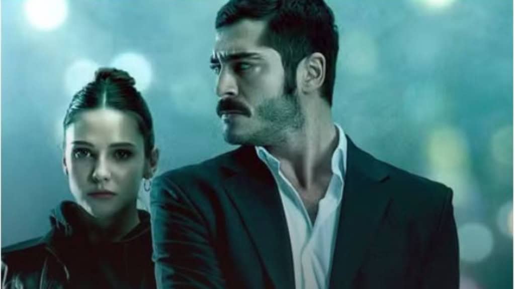 Dois personagens se encaram de perto com expressões sérias; um de terno observa o outro, que usa jaqueta escura, contra um fundo azul desfocado.