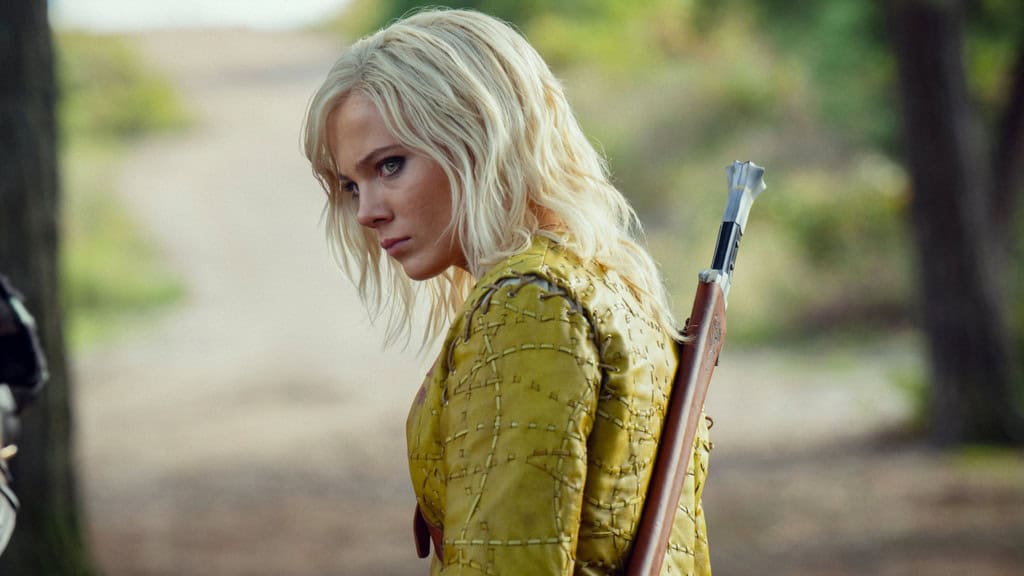Ciri, personagem de The Witcher, aparece com cabelos loiros e expressão séria, vestindo jaqueta amarela e carregando uma espada nas costas, em cena da 4ª temporada da série da Netflix