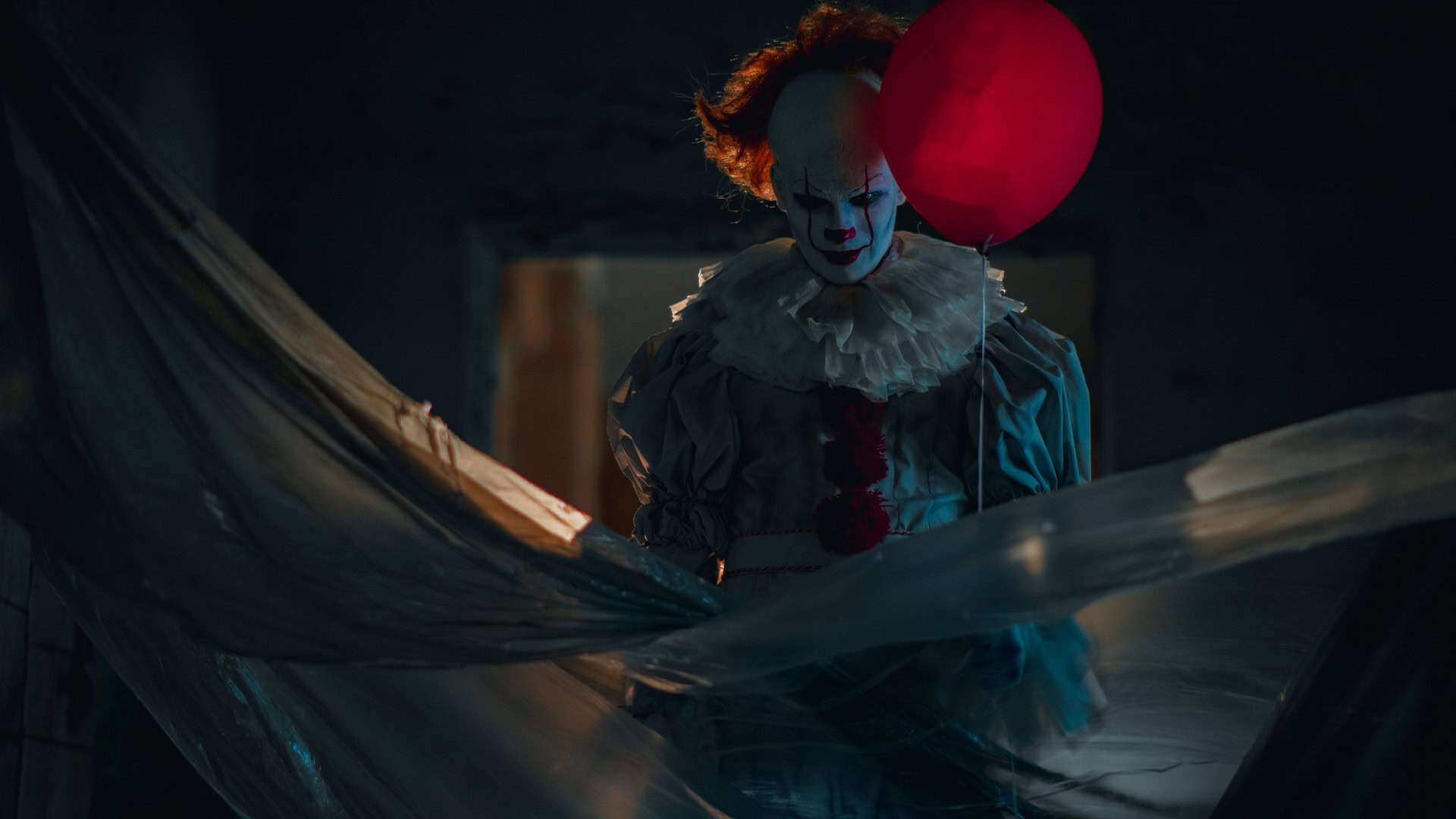 Um palhaço assustador, com rosto branco e maquiagem vermelha, segura um balão vermelho. Ele veste uma roupa antiga com babados e pompons vermelhos. Está parcialmente escondido atrás de plásticos transparentes, em um lugar escuro e abandonado. A cena transmite medo, mistério e tensão.