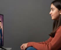 Uma pessoa sentada no chão observa atentamente a TV, que mostra sete figuras em pé sob um céu dramático, sugerindo cena de série ou filme.