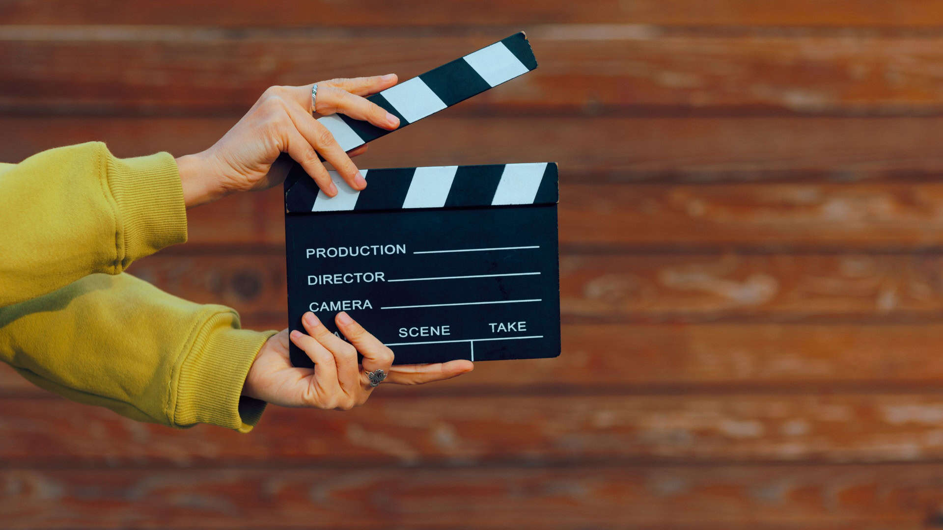 Uma mão com anéis segura uma claquete de cinema preta e branca diante de uma parede de madeira.