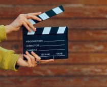 Uma mão com anéis segura uma claquete de cinema preta e branca diante de uma parede de madeira.