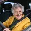 Idosa sorridente dirige um carro, usando cinto de segurança e óculos, transmitindo autonomia e bem-estar.