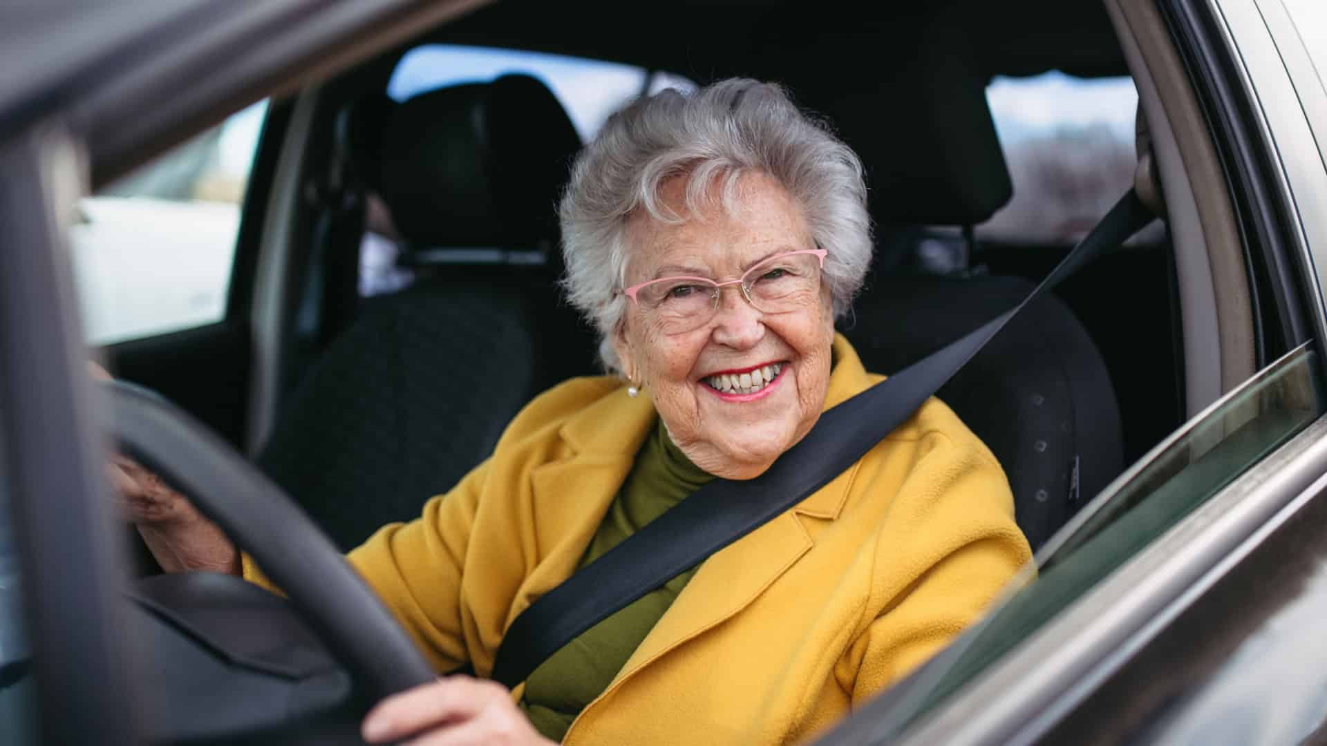 Idosa sorridente dirige um carro, usando cinto de segurança e óculos, transmitindo autonomia e bem-estar.
