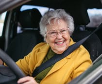 Idosa sorridente dirige um carro, usando cinto de segurança e óculos, transmitindo autonomia e bem-estar.