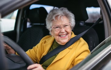Idosa sorridente dirige um carro, usando cinto de segurança e óculos, transmitindo autonomia e bem-estar.